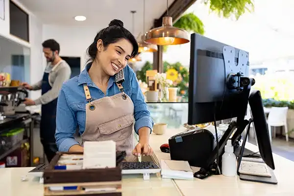 A cheerful barista wearing an apron multitasks behind the counter, speaking on the phone while using a computer at a coffee shop.