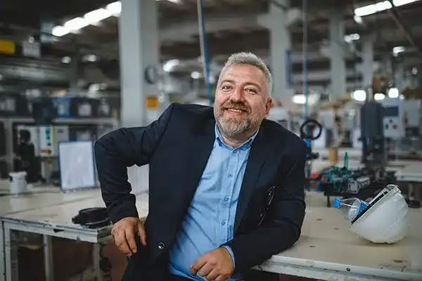 A smiling man in a business suit stands confidently in an industrial setting, leaning against a workbench in a factory.